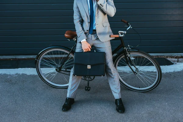 Обрізаний вид бізнесмена тримає портфель, стоячи біля велосипеда і будуючи на відкритому повітрі — стокове фото