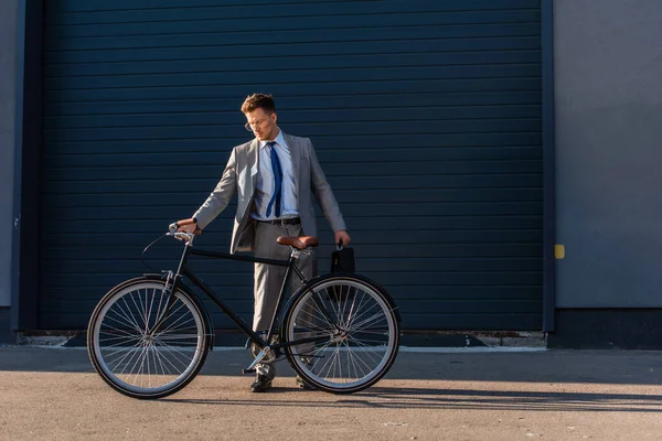 Empresário em uso formal segurando pasta perto de bicicleta e construção ao ar livre — Fotografia de Stock