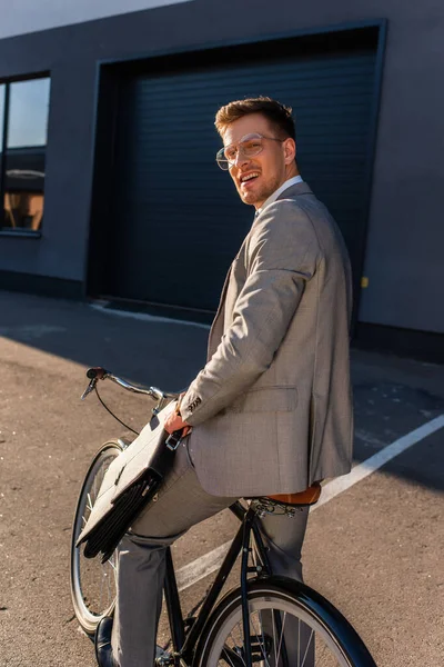 Jeune homme d'affaires souriant à la caméra tout en tenant la mallette sur le vélo à l'extérieur — Photo de stock