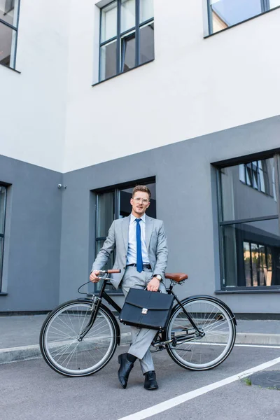 Uomo d'affari in occhiali e tuta che tiene la valigetta vicino alla bici e all'edificio — Foto stock