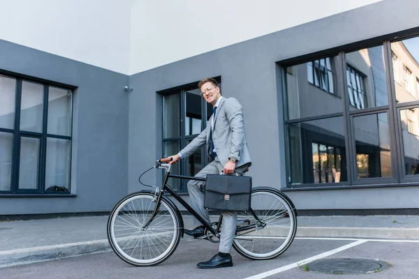 Businessman smiling at camera while holding briefcase and riding bike outdoors — Stock Photo