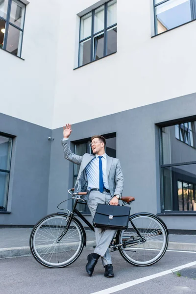 Веселий бізнесмен махає рукою, тримаючи портфель біля велосипеда на відкритому повітрі — стокове фото