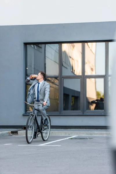 Joven hombre de negocios bebiendo café para ir en bicicleta cerca del edificio - foto de stock