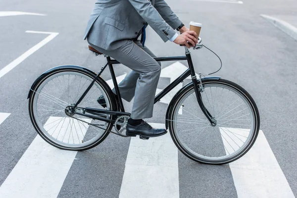 Обрезанный вид бизнесмена, держащего кофе на вынос во время езды на велосипеде по пешеходной дорожке — стоковое фото