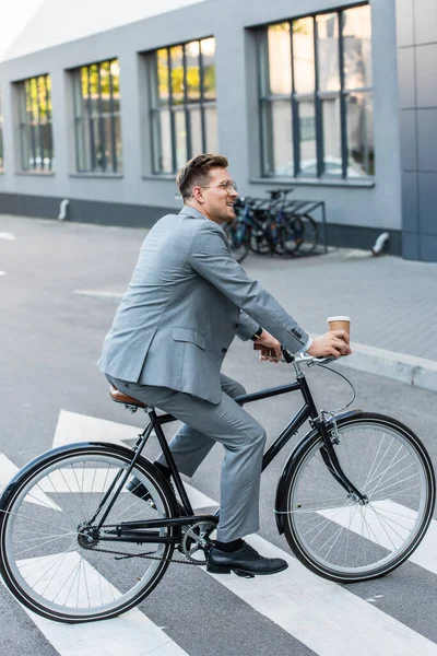 Smiling businessman with coffee to go riding bicycle on crosswalk outdoors — Stock Photo