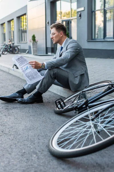 Young businessman reading newspaper while sitting on walkway near bike on blurred foreground — Stock Photo