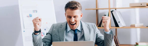 Positive businessman showing yeah gesture near laptop on blurred foreground, banner — Stock Photo