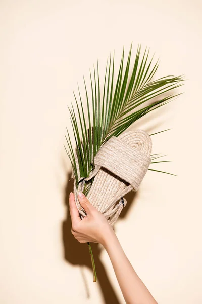 Обрезанный вид женщины, держащей летние плетеные сандалии и пальмовый лист на бежевом фоне — стоковое фото