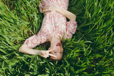 Genç eğlence alanında güneşli havalarda parlak yeşil arka plan üzerinde dinlenme zafer işaret gösteren çim yatan hafif desenli elbiseli güzel kadın çok memnun. Bahar doğa. Yaşam tarzı kavramı