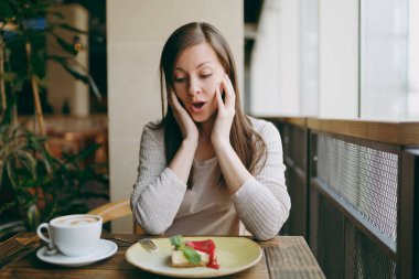 Çekici genç kadın tek başına yakınındaki kafede masada büyük pencere kapuçino, bardak ile oturmak kek, restoranda serbest zaman sırasında rahatlatıcı. Geri kalan kafede olan genç kadın. Yaşam tarzı kavramı