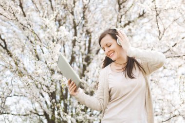 Genç güzel kadın hafif rahat kıyafetler tablet pc bilgisayar şehir bahçe veya park çiçek açan ağaç arka planda tutan müzik dinlerken kulaklık ile rahat. Bahar çiçekleri. Yaşam tarzı kavramı