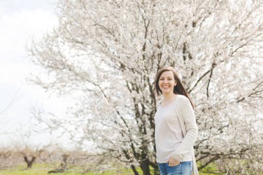 Genç mutlu gülümseyen güzel kadın elleri ceplerinde şehir bahçe veya park çiçek açan ağaç arka plan üzerinde ayakta tutmak hafif rahat kıyafetler içinde. Bahar doğa, çiçekler. Yaşam tarzı, eğlence kavramı