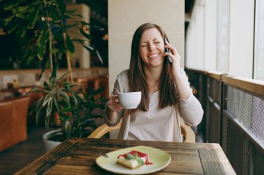 Yalnız fincan kapuçino, kafede masada oturan genç kadın kek, restoranda serbest zaman sırasında rahatlatıcı. Dinlenme kafede olan genç kadın cep telefonda konuşurken. Yaşam tarzı kavramı