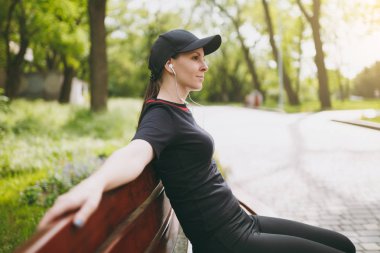 Genç atletik oldukça esmer kız siyah üniforma giyen rahat ve şehir park açık havada bankta müzik dinleme eğitimi egzersiz sonra dinlenme kulaklık ile kap. Fitness, sağlıklı yaşam konsepti