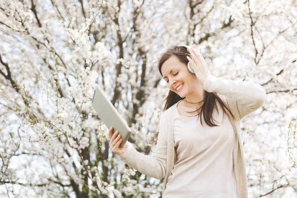 Young relaxed beautiful woman in light casual clothes with headphones listening music holding tablet pc computer in city garden or park on blooming tree background. Spring flowers. Lifestyle concept