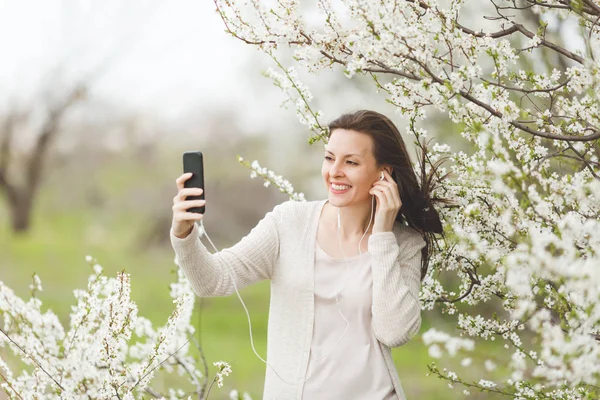 Selfie cep telefonu şehir Bahçe veya park çiçek açan ağaç arka plan üzerinde yapıyor müzik dinlerken kulaklık ile hafif rahat kıyafetler genç mutlu güzel kadın. Bahar çiçekleri. Yaşam tarzı kavramı
