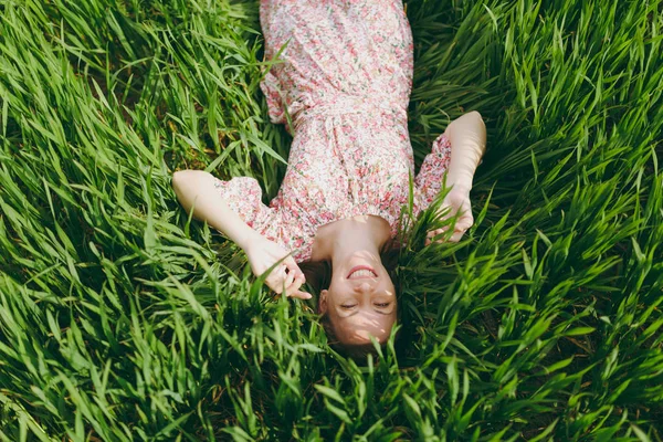 Parlak yeşil arka plan üzerinde alanında güneşli havalarda dinlenme seyir eller yaymak çim üzerinde yalan hafif desenli elbiseli genç gülümseyen güzel kadın. Bahar doğa. Yaşam tarzı, eğlence kavramı