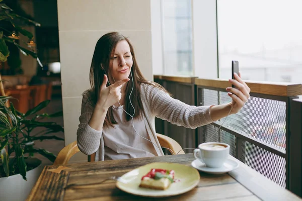 Yalnız fincan kapuçino, kafede masada oturan genç kadın kek, restoranda serbest zaman sırasında rahatlatıcı. Genç selfie cep telefonu, yapıyor kadın cafede bekletin. Yaşam tarzı kavramı