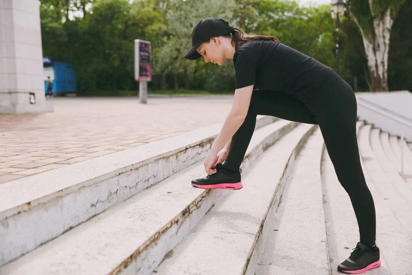 Genç atletik güzel esmer kız siyah üniforma ve cap city Park açık havada merdivenlerde bağlama ayakkabı bağcığı çalıştırmadan önce ısınma spor egzersizleri, yapıyor. Fitness, sağlıklı yaşam konsepti
