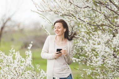 Genç gülümseyen güzel kadın hafif rahat kıyafetler içinde cep telefonu şehir bahçe veya park çiçek açan ağaç arka planda tutan müzik dinlerken kulaklık ile rahat. Bahar çiçekleri. Yaşam tarzı kavramı