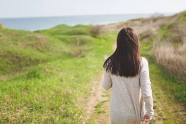 Görünüm genç esmer kadın tarlada su olarak güneşli havalarda yol boyunca yeşil arka plan üzerinde yürüyen ışık rahat kıyafetler içinde geri. Güzel manzara. Yaşam tarzı, eğlence kavramı