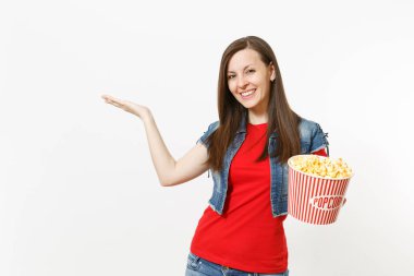 Film film izlerken, kova patlamış mısır holding, el bir kenara beyaz arka plan üzerinde izole boşaltmak üzerinde işaret eden rahat kıyafetler içinde genç gülümseyen güzel kadın portresi. Sinema kavramı duygular