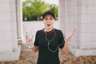 Genç atletik siyah üniforma giyen güzel esmer kız şok ve şişe su ile holding, eller şehir park açık havada yayılan müzik dinlerken kulaklık ile kap. Fitness, sağlıklı yaşam tarzı