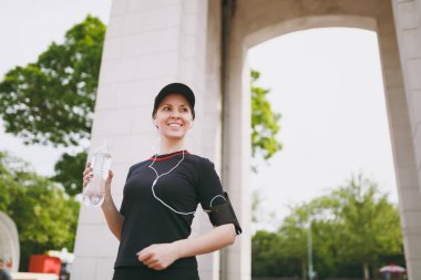 Genç atletik gülümseyen güzel esmer kız siyah üniforma ve eğitim, dinleme müzik Şehir Park açık havada sırasında şişe su ile tutarak kulaklık ile kap. Fitness, sağlıklı yaşam tarzı