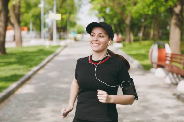 Atletik esmer kız siyah üniforma giyen ve kulaklık ile kap gülümseyen genç spor yapıyor, çalıştıran, koşu, city Park açık havada yolda müzik eğitimi. Fitness, sağlıklı yaşam konsepti
