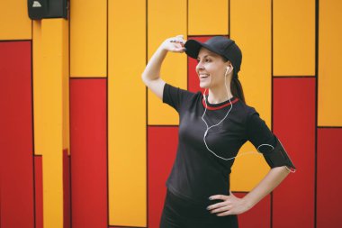 El cap yakınındaki açık havada parlak arka planda tutan genç gülümseyen atletik güzel esmer kız siyah üniforma ve bir yana ayakta, seyir kulaklıklar ile kap. Fitness, sağlıklı yaşam konsepti