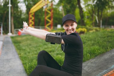 Siyah üniforma uzanan eller, dinlenme ve önce veya sonra şehir park açık havada çalışan, eğitim oturan müzik dinlerken kulaklık ile genç atletik gülümseyen kız. Fitness, sağlıklı yaşam tarzı