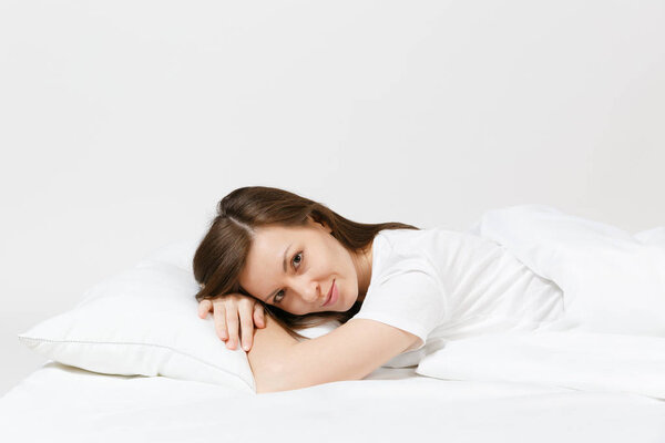 Calm young brunette woman lying in bed with white sheet, pillow, blanket on white background. Smiling beauty female spending time in room. Rest, relax, good mood concept. Copy space for advertisement