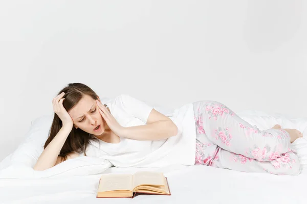 Young dissatisfied woman lying in bed with white sheet, pillow, blanket on white background. Female spending time in room, reading book, enjoying of rest. Relax, good mood concept. Place for text