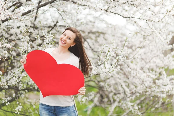 Young happy beautiful woman in light casual clothes holding big red heart standing in city garden or park on blooming tree background. Copy space for advertisement. Spring flowers. Lifestyle concept