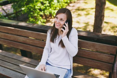 Cep telefonu üzerinde konuşurken hafif rahat kıyafetler genç mutlu kadın. Modern dizüstü pc bilgisayar açık havada doğa üzerinde sokakta çalışan bankta oturan kadın. Mobil ofis. Serbest iş kavramı