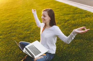Genç öğrenci rahat kıyafetler içinde kadın sakin. Kadın oturuyordu, rahatlatıcı, yoga hareketle eller. Dizüstü pc bilgisayar yeşil çimenlerin üzerinde açık havada Güneş çim zemin. Mobil ofis. Serbest iş kavramı