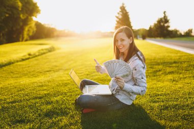 Kadın holding bohça-in dolar nakit para. Dizüstü pc bilgisayar Şehir Park açık havada Güneş çim yeşil çim üzerinde çalıştığı yer, çimenlerin üzerinde oturan bir bayan. Mobil ofis. Serbest iş kavramı