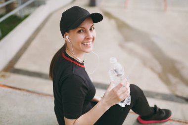 Siyah üniformalı genç atletik gülümseyen kız kap önce veya sonra şehir Park açık havada çalışan, eğitim oturan su ile şişe tutan müzik dinlerken kulaklık ile. Fitness, sağlıklı yaşam tarzı