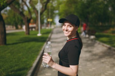 Genç gülümseyen güzel esmer kız siyah üniforma giyen ve ayakta açık havada şehir parkında çalıştırmadan önce eğitim sırasında su kap holding şişeyle atletik. Fitness, sağlıklı yaşam konsepti