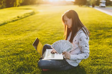 Kadın holding bohça-in dolar nakit para. Dizüstü pc bilgisayar Şehir Park açık havada Güneş çim yeşil çim üzerinde çalıştığı yer, çimenlerin üzerinde oturan bir bayan. Mobil ofis. Serbest iş kavramı