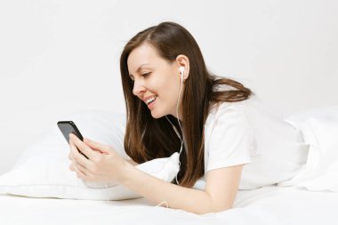 Beyaz çarşaf, yastık, battaniye beyaz arka plan üzerinde ile yatakta yalan sakin genç kadın. Güzellik kadın dinleme müzik kulaklık içinde hareket eden telefon, odasında vakit. Dinlenmek, dinlenmek iyi ruh kavramı