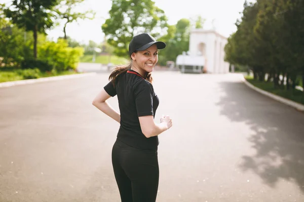 stock image Young athletic smiling beautiful brunette girl in black uniform and cap training, doing sport exercises and running, looking back on path in city park outdoors. Fitness, healthy lifestyle concept.