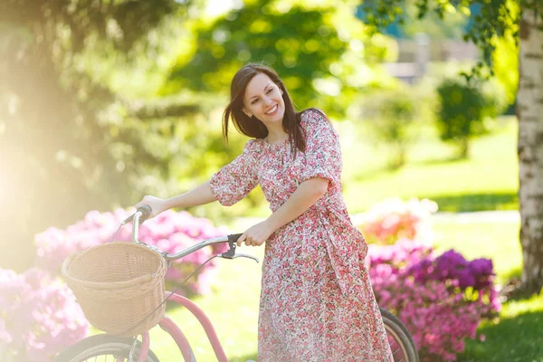 Portrait of trendy young woman in long pink floral dress stop to riding on vintage bike with basket for purchases, flowers background outdoors. Pretty female recreation time in spring or summer park