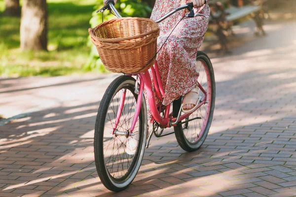 Close up cropped photo of young woman in long pink floral dress riding on vintage bike with basket for purchases, food or flowers outdoors, wheels, female recreation time in spring or summer park