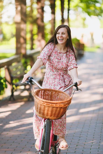 Portrait of trendy young woman in long pink floral dress riding on alley on vintage bike with basket for purchases, food or flowers outdoors, gorgeous female recreation time in spring or summer park