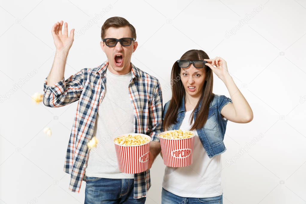 Young irritated couple, woman and man in 3d glasses and casual clothes watching movie film on date holding buckets of popcorn, popping popcorn isolated on white background. Emotions in cinema concept