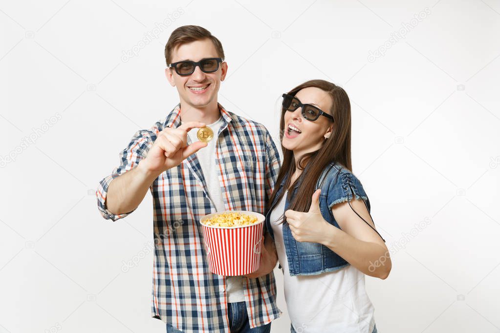 Young happy smiling couple, woman and man in 3d glasses and casual clothes watching movie film on date, holding bucket of popcorn and bitcoin isolated on white background. Emotions in cinema concept