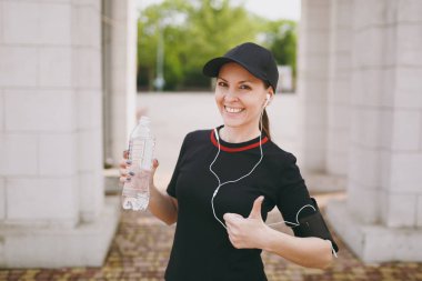 Genç atletik gülümseyen güzel esmer kız siyah üniforma giyen ve şişe su, Şehir Parkı açık başparmak gösterilmesini müzik dinleme ile tutarak kulaklık ile kap. Fitness, sağlıklı yaşam tarzı