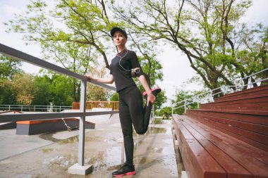 Genç atletik güzel esmer kız siyah üniformalı, ısınma-up şehir park açık havada spor germe egzersizleri yapıyor müzik dinlerken kulaklık ile kap konsantre. Fitness, sağlıklı yaşam tarzı