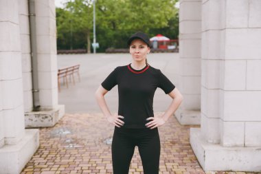 Genç gülümseyen atletik güzel esmer kız siyah üniforma ve spor egzersizleri, çalıştırmadan önce ısınma yapıyor cap city park açık havada duran. Fitness, sağlıklı yaşam konsepti